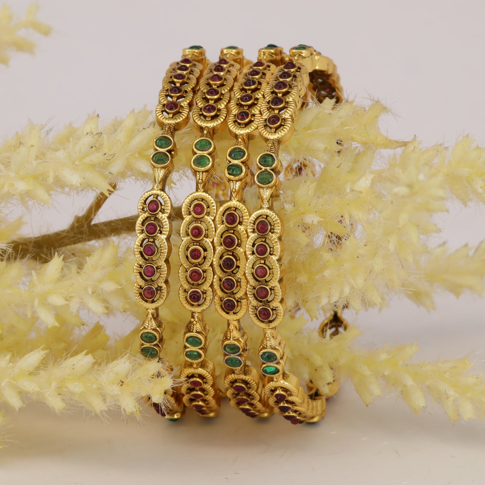 Exquisite gold bangles featuring layered floral patterns with ruby-red and emerald-green stones, crafted in a rich traditional style ideal for weddings and festive celebrations.